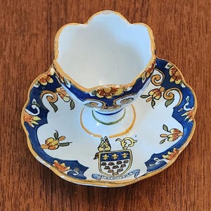 May include: A ceramic egg cup with a matching saucer featuring a scalloped edge, painted with blue, yellow, and orange floral patterns. The saucer displays a central crest with a bishop's mitre and the text Mt-St-Michel in blue lettering.