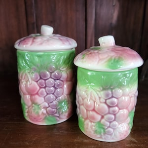 May include: Two ceramic canisters with lids, featuring a green and pink fruit design. The canisters are decorated with grapes, strawberries, and cherries.