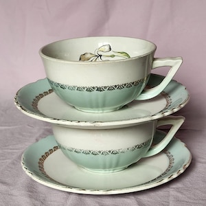 Peut inclure: Deux tasses à thé et soucoupes en céramique empilées présentant un coloris vert menthe pâle et blanc. Chaque pièce est ornée d'une bande argentée métallique avec un motif de feuilles répétitif. L'intérieur de la tasse supérieure montre un motif floral.