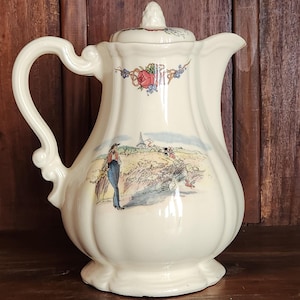Può includere: Una brocca in ceramica color crema con coperchio, caratterizzata da un corpo smerlato e un manico. La superficie è decorata con un'illustrazione colorata di una persona in un campo vicino a una chiesa, accentuata da un motivo floreale sul bordo.