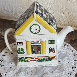Puede incluir: Una tetera de cerámica con forma de estación de tren con techo de tejas, un reloj y pilares amarillos. El frente presenta el texto Primrose Junction, OXO y a railway station teapot. Incluye detalles pintados de personas, un gato y flores.