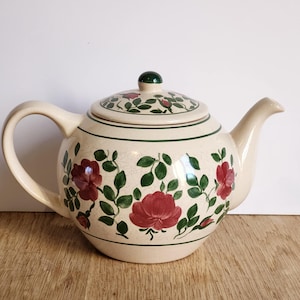 Peut inclure: Théière en céramique crème avec un couvercle, une anse et un bec verseur. La théière est ornée de roses rouges et de feuilles vertes. Une bande verte entoure la base et le couvercle. Le couvercle a un bouton vert.