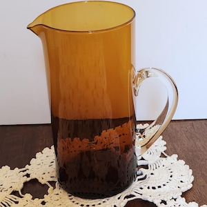 May include: A cylindrical glass pitcher featuring a deep amber colour that transitions to a darker brown at the base. The vessel has a clear, curved glass handle attached to the side and a small, integrated pouring spout at the top rim.