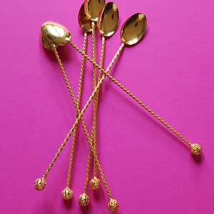 Pode incluir: Um conjunto de seis colheres douradas com cabos longos e torcidos e extremidades decorativas em forma de bola. As colheres têm conchas ovais. As colheres estão dispostas sobre um fundo magenta vibrante.