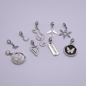 Könnte beinhalten: Eine Sammlung von silberfarbenen Anhängern mit verschiedenen Designs, darunter ein Schmetterling, ein Halbmond, eine Katze, ein Flugzeug, ein Schneeflocke, ein Dreieck und ein Anhänger mit dem Wort „ENOUGH“. Die Anhänger sind wahrscheinlich für die Schmuckherstellung oder als dekorative Elemente gedacht.