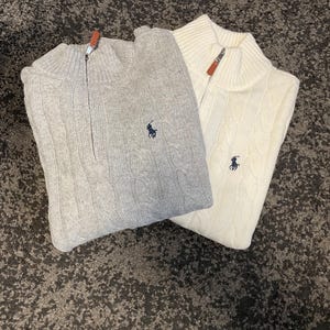 Puede incluir: Dos suéteres de punto trenzado, uno gris claro y otro blanco roto, están doblados sobre una superficie texturizada. Cada suéter tiene un cuello con cremallera y un pequeño logotipo azul marino en el pecho. Las cremalleras tienen tiradores naranjas.