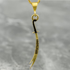 May include: A gold pendant necklace featuring a detailed, curved sword design with black accents. The sword is inscribed with Arabic calligraphy.