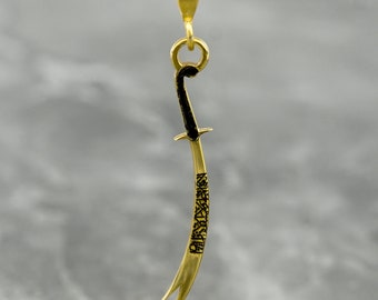 May include: A gold pendant necklace featuring a detailed, curved sword design with black accents. The sword is inscribed with Arabic calligraphy.