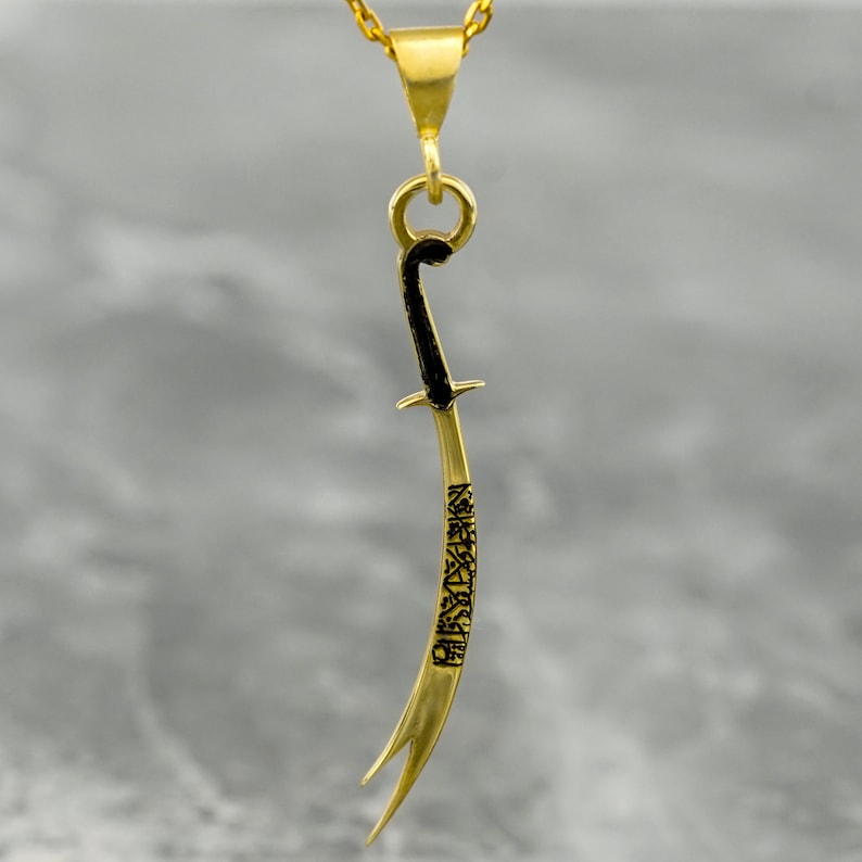 May include: A gold pendant necklace featuring a detailed, curved sword design with black accents. The sword is inscribed with Arabic calligraphy.