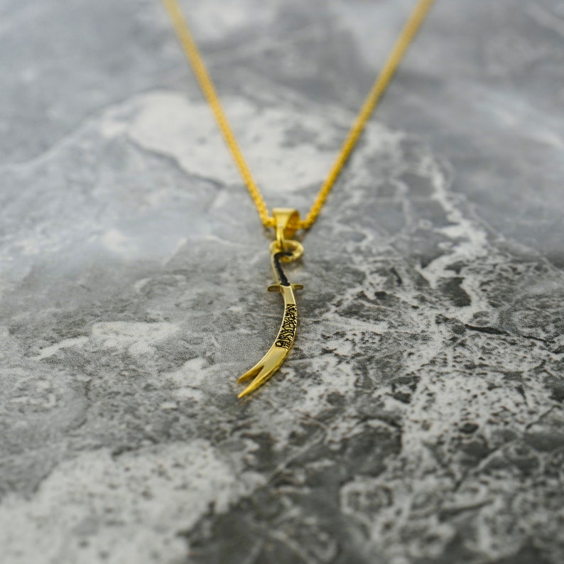 May include: A gold chain necklace with a pendant shaped like a curved sword. The sword has Arabic writing on it.