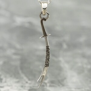 May include: Silver pendant necklace featuring a detailed, curved sword design with Arabic script along the blade.