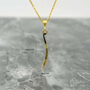 May include: Gold pendant necklace featuring a black sword with Arabic calligraphy. The pendant is 1.41 inches tall and 0.01 inches wide.