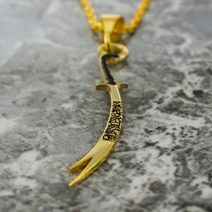 May include: A gold-colored pendant necklace featuring a sword design with intricate details. The sword is curved and has Arabic script engraved on it.