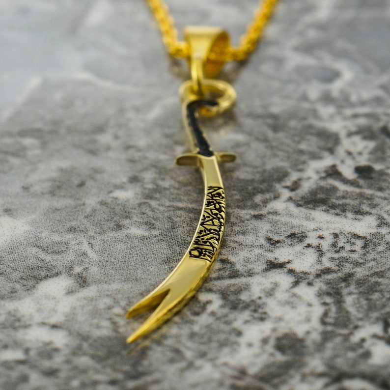 May include: A gold-colored pendant necklace featuring a sword design with intricate details. The sword is curved and has Arabic script engraved on it.