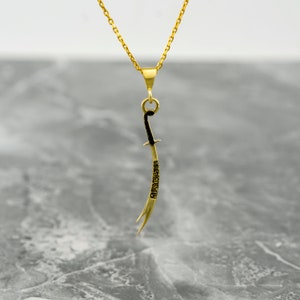 May include: A gold chain necklace with a pendant shaped like a curved sword. The sword is black and has Arabic script engraved on it.