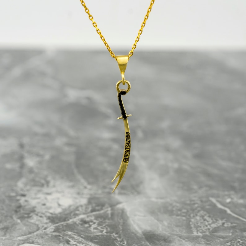 May include: A gold chain necklace with a pendant shaped like a curved sword. The sword is black and has Arabic script engraved on it.