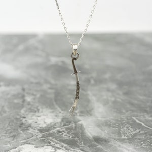 May include: A silver necklace with a pendant shaped like a curved sword. The sword has Arabic script engraved on it.