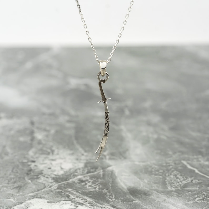 May include: A silver necklace with a pendant shaped like a curved sword. The sword has Arabic script engraved on it.