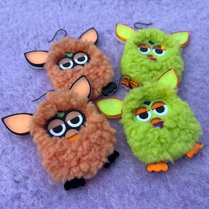 May include: Three fuzzy, colorful, plush toy earrings. Two are orange and one is green. Each earring has a face with large eyes and a small mouth. The earrings are hanging from a light purple background.
