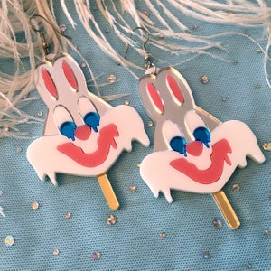 May include: A pair of white and pink bunny rabbit face earrings with blue eyes and a dripping red smile. The earrings are on a stick and have gold-colored hooks.