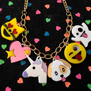 May include: A gold-colored chain necklace with various emoji-style charms. Charms include a yellow cat with heart eyes, a pink heart with stars, a white unicorn, a dog, a yellow emoji with a tongue sticking out, and a white ghost.