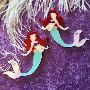 May include: A pair of mermaid-themed earrings with red hair, a white bodice, and a turquoise tail. The earrings are made of iridescent acrylic and have silver hooks.