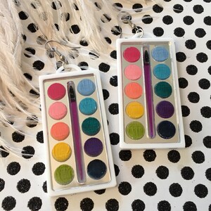 Puede incluir: Un par de pendientes de paleta de pintura de acuarela con un dije de pincel plateado. Las paletas son blancas con un borde plateado y tienen 10 pocillos de pintura de colores.
