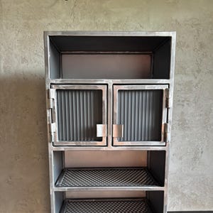 Könnte beinhalten: Ein Metallschrank mit rechteckigem Rahmen. Er verfügt über zwei Glastüren und zwei Drahtregale. Der Schrank ist grau lackiert und hat ein minimalistisches Design. Der Schrank ist für die Aufbewahrung geeignet.
