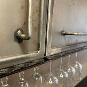 May include: A metal cabinet with a silver finish and a metal handle. The cabinet is mounted above a wine glass rack with several wine glasses hanging from it.