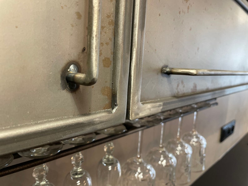 May include: A metal cabinet with a silver finish and a metal handle. The cabinet is mounted above a wine glass rack with several wine glasses hanging from it.