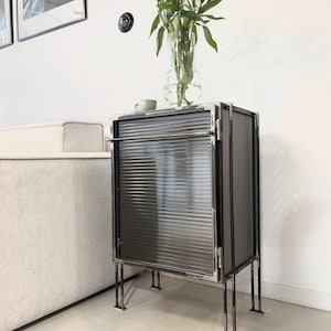 May include: A black metal cabinet with a glass door and a metal frame. The cabinet has a slatted design on the door and sits on four thin metal legs. A vase with white flowers sits on top of the cabinet.