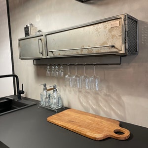 May include: A metal cabinet with a distressed finish and a wire mesh door. The cabinet is mounted on a wall above a black countertop with a wooden cutting board and a metal wine glass rack. The cabinet has a small metal tag with the word "MIGHTY" on it.