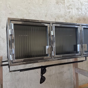 Può includere: Un mobile metallico con tre ante in vetro. Il mobile ha un'estetica scura e industriale con una barra nera sottostante, che tiene un paio di occhiali da sole neri. Il mobile è montato su una parete chiara.