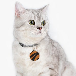 May include: A silver tabby cat wearing a black collar with a round orange and black pet tag. The cat has green eyes and is looking to the side against a white background. The tag has a tiger stripe design.