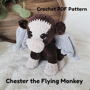 May include: A handmade crochet plushie of a monkey with gray wings, white face, and brown body. The text "Crochet PDF Pattern" and "Chester the Flying Monkey" are displayed. The plushie is sitting on a white, textured surface.