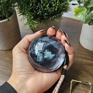Può includere: Una sfera di vetro trasparente con un'immagine in bianco e nero all'interno, tenuta in mano. La sfera ha un cordino a fantasia rosa e nero attaccato, insieme a un anello quadrato color oro.