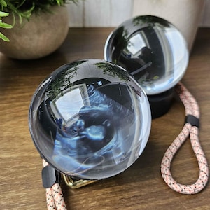 May include: Two clear crystal balls with black bases and patterned lanyards rest on a wooden surface. Green plants in gray pots are in the background. The crystal balls reflect the surrounding environment.
