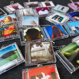 May include: A collection of miniature CD case keychains. Each clear plastic case holds a small album cover image. The album art features various colors and designs, including portraits and landscapes. Some cases have a silver chain attached.