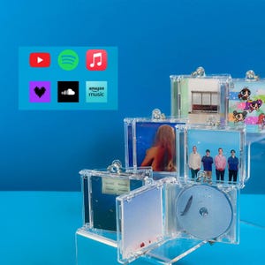 Könnte beinhalten: Ein klarer CD-Display-Ständer aus Acryl mit mehreren CD-Hüllen, von denen einige Albumcover zeigen. Der Ständer steht auf einer blauen Oberfläche, mit App-Symbolen für YouTube, Spotify und Amazon Music im Hintergrund. Die CDs sind sichtbar.