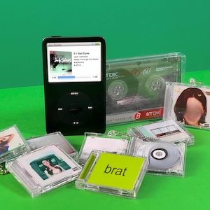 May include: A black iPod with a screen displaying a Jack Johnson song, surrounded by miniature CD cases and a cassette tape. The CD cases have various album art, and one reads "brat" in green. The background is a vibrant green.