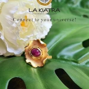Peut inclure: Une bague dorée avec un design floral, ornée d'une pierre précieuse ovale rouge. La bague est présentée sur une feuille verte, avec une fleur blanche en arrière-plan. Le texte indique "LA KIATRA HANDMADE JEWELLERY" et "Connect to your universe!"