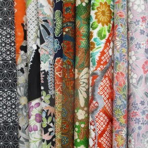May include: A collection of vertical fabric rolls featuring diverse patterns including black and white geometric shapes, orange floral motifs, dark blue botanical prints, intricate red and gold designs, and light pink floral arrangements.