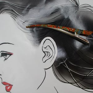 May include: A silver-toned metal hair clip featuring a curved, elongated design with a vibrant, multi-coloured pattern in shades of orange, green, and blue. The clip has a smooth, polished finish on the underside and a decorative top surface.