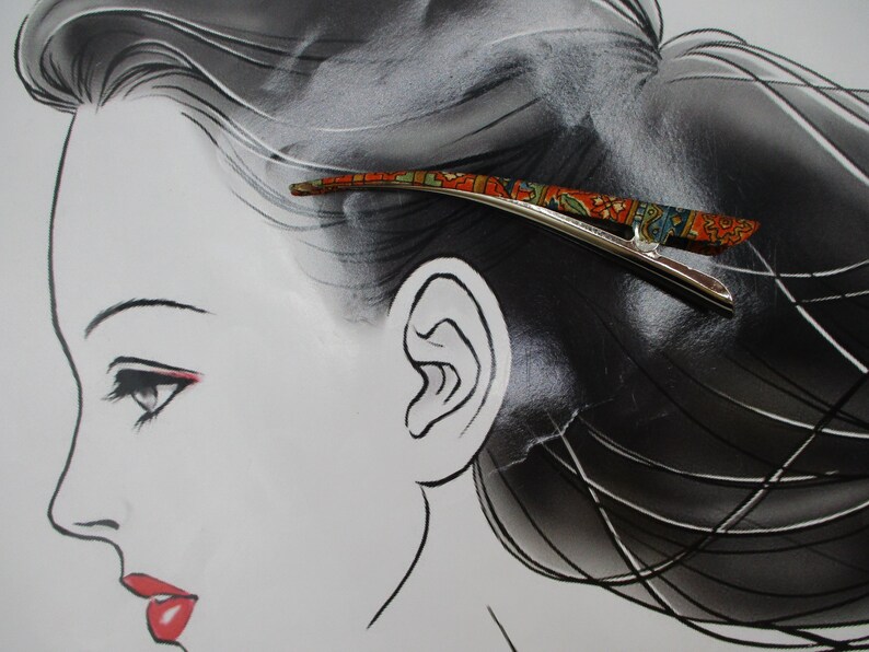 May include: A silver-toned metal hair clip featuring a curved, elongated design with a vibrant, multi-coloured pattern in shades of orange, green, and blue. The clip has a smooth, polished finish on the underside and a decorative top surface.