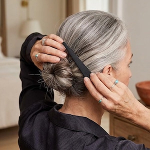 May include: A black hair comb is being used to style a bun. The person is wearing a black top and rings with turquoise stones. The hair is gray and styled in a low bun.