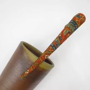 May include: A long, slender, tapered hair stick featuring a vibrant orange base with intricate blue, green, and white floral and geometric patterns, resting inside a smooth, matte brown conical cup with a wide, open rim.
