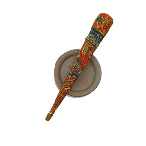 May include: A tapered hair stick featuring a vibrant orange base colour with intricate, multi-coloured floral and geometric patterns in shades of blue, green, and yellow, resting diagonally across a plain, circular, light grey disc-shaped base.
