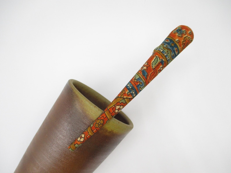 May include: A long, slender, tapered hair stick featuring a vibrant orange base with intricate blue, green, and white floral and geometric patterns, resting inside a smooth, matte brown conical cup with a wide, open rim.