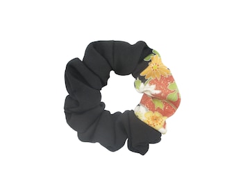 May include: A fabric hair scrunchie featuring a solid black section and a contrasting section with a floral pattern in shades of orange, yellow, and green. The accessory has a gathered, elasticized design that creates a circular, ruffled appearance.