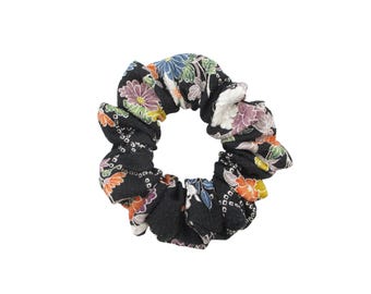 May include: A black fabric hair scrunchie featuring a repeating floral pattern in shades of purple, orange, white, and green. The accessory has a gathered, ruffled texture throughout the circular band, creating a voluminous and soft appearance.